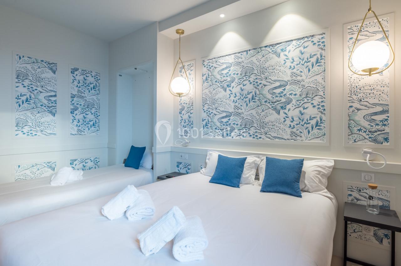 Chambre lumineuse avec lit double, lit simple, décor mural bleu et blanc, et luminaires suspendus modernes.