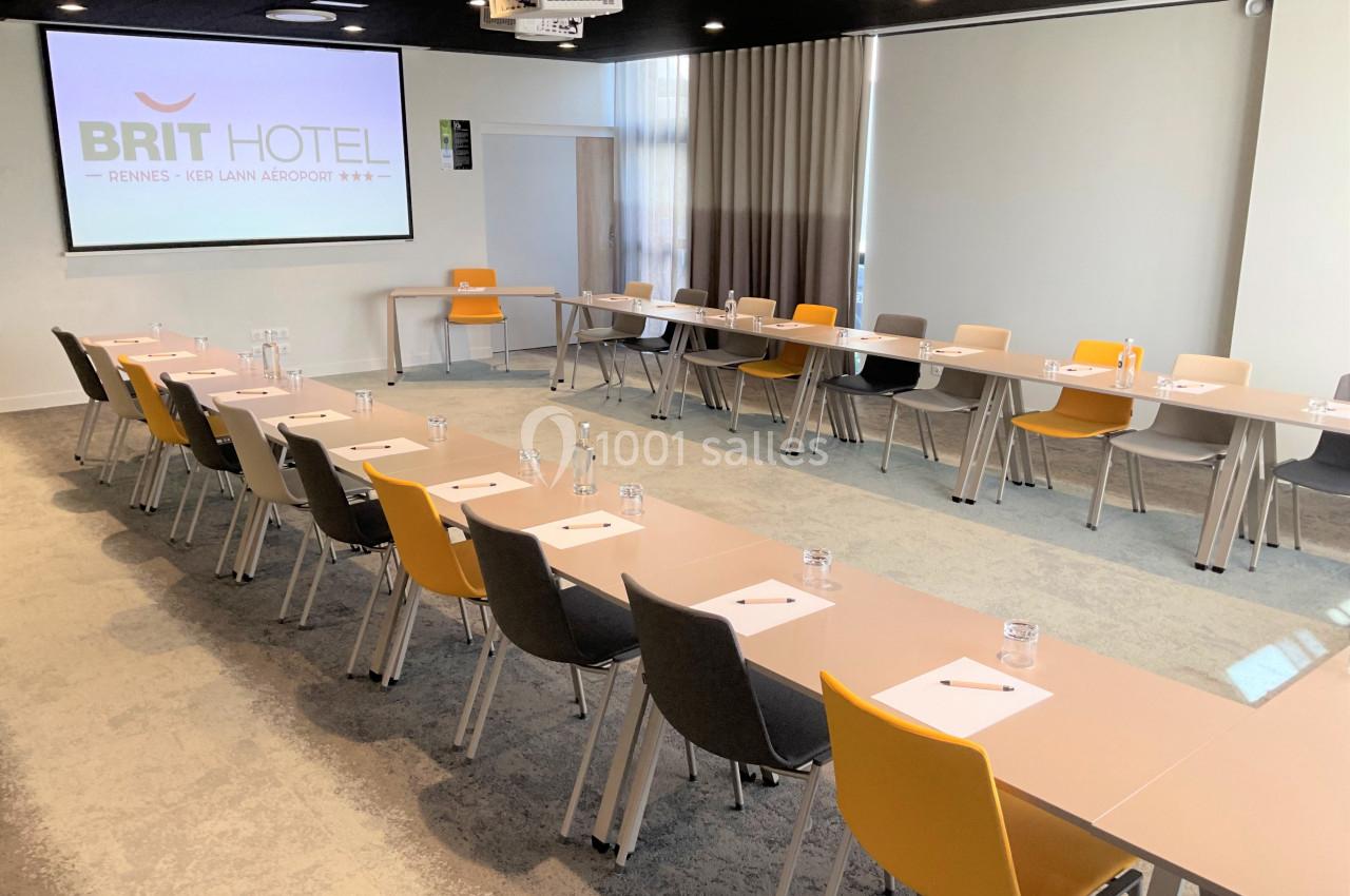 Salle de réunion lumineuse avec tables disposées en U, chaises colorées et écran de projection allumé.