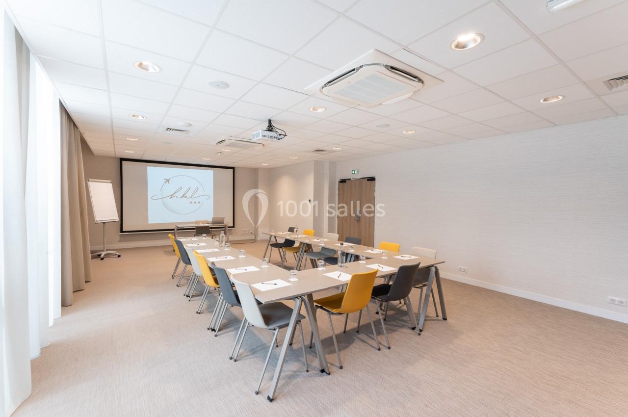 Salle de réunion lumineuse avec tables disposées en U, chaises colorées, écran de projection et paperboard.