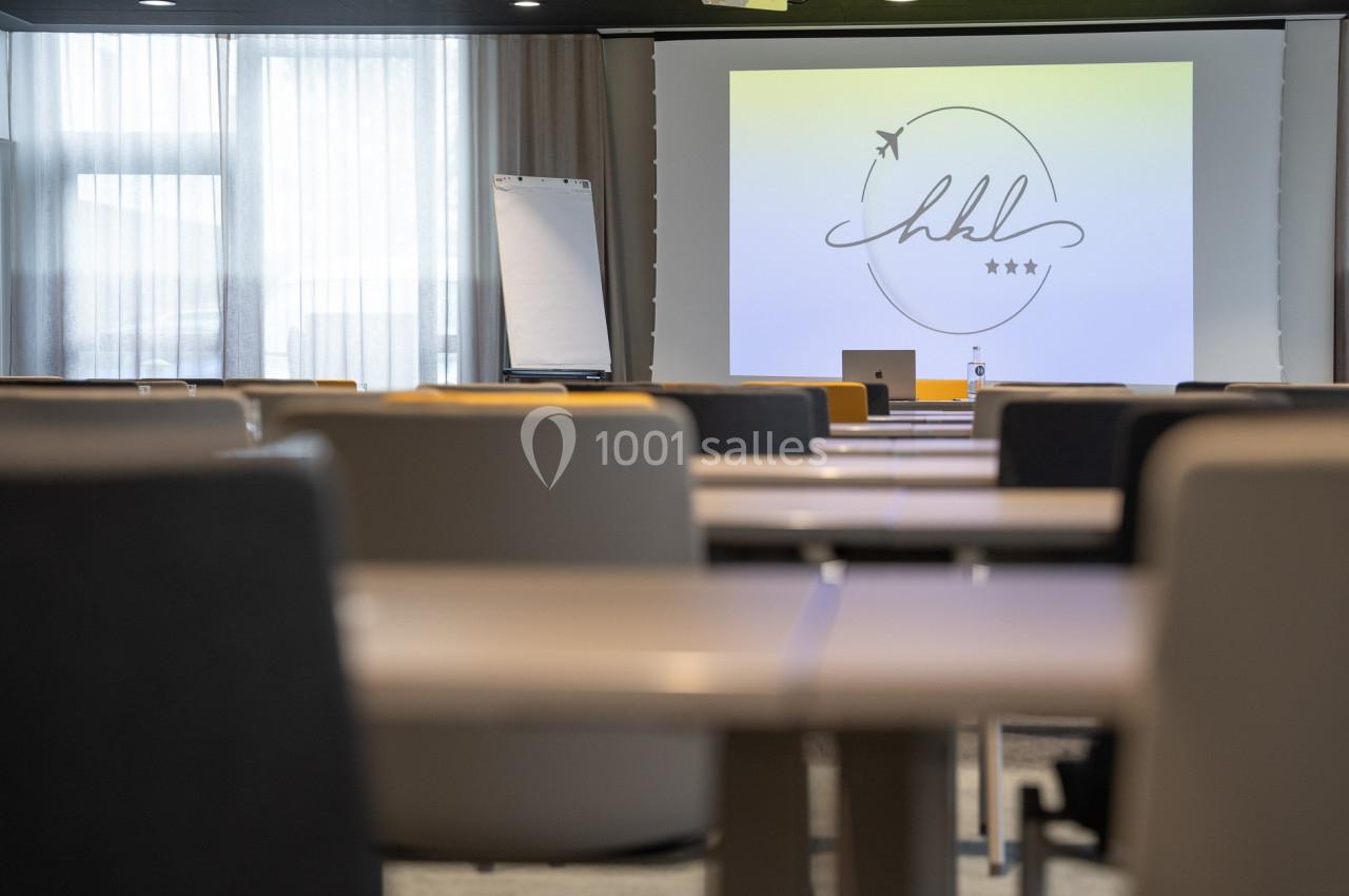 Salle de conférence lumineuse avec des rangées de tables, un écran de projection et un tableau blanc en arrière-plan.