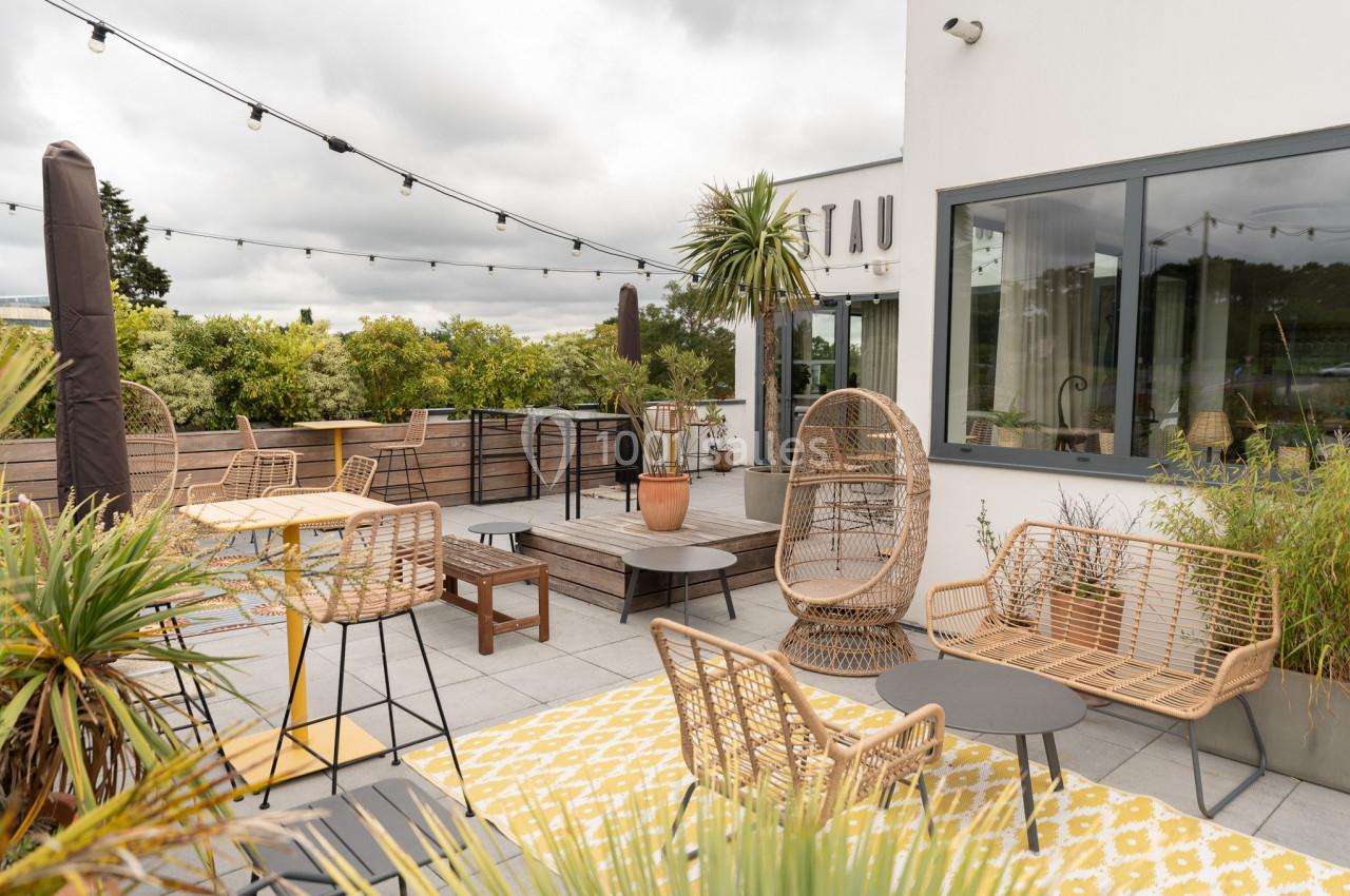 Terrasse aménagée avec mobilier en rotin, tables, plantes et guirlandes lumineuses, entourée de verdure.
