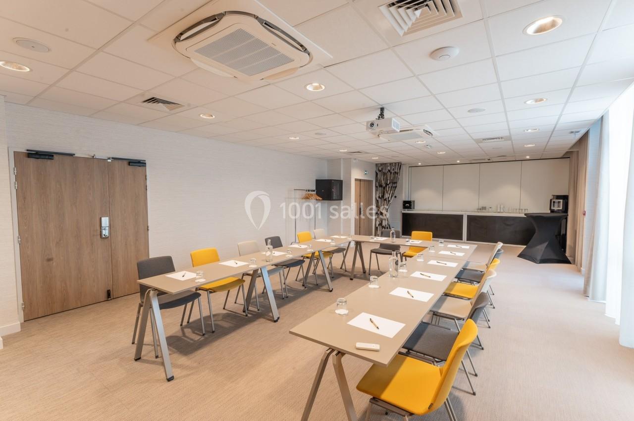 Salle de réunion lumineuse avec tables disposées en U, chaises jaunes et grises, et équipements audiovisuels.