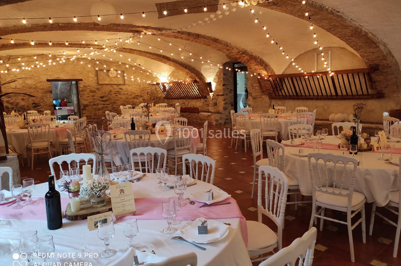 Salle de réception décorée avec des tables dressées, nappes blanches et roses, éclairée par des guirlandes lumineuses.