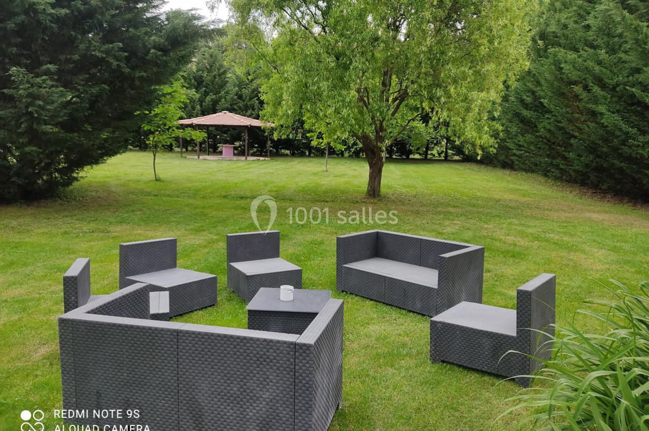 Mobilier de jardin en résine tressée disposé sur une pelouse, avec un kiosque en arrière-plan entouré d'arbres.