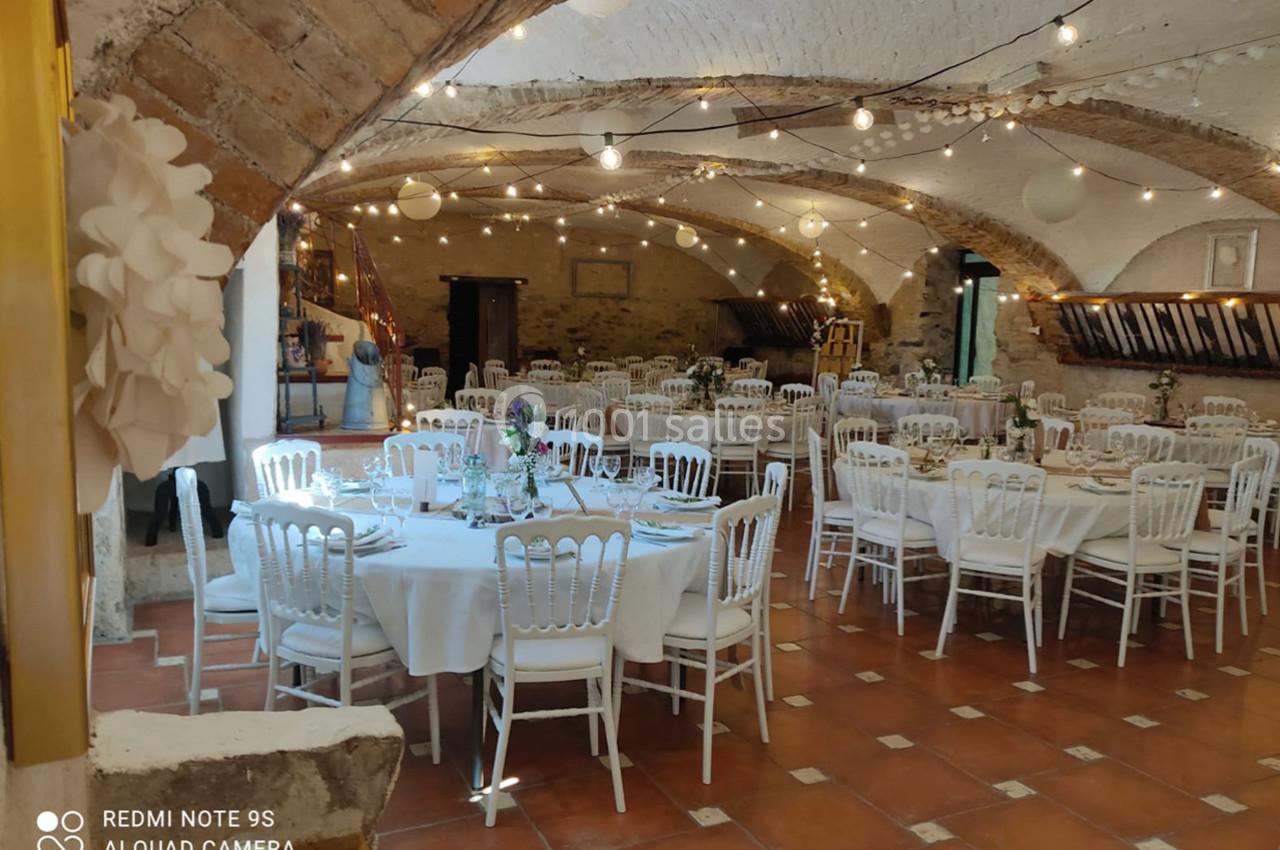 Salle voûtée décorée pour un événement, avec tables rondes dressées, chaises blanches et guirlandes lumineuses suspendues.
