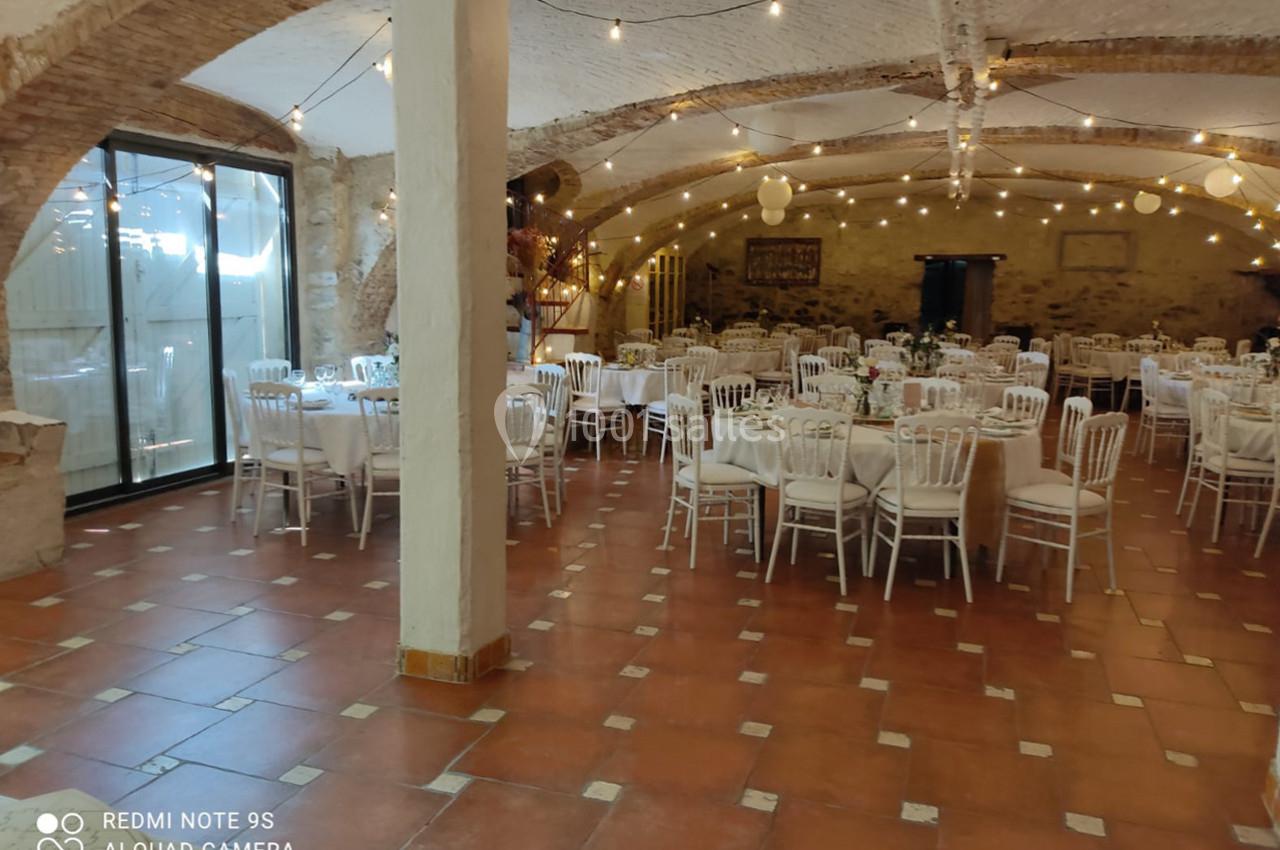 Salle de réception avec tables rondes dressées, chaises blanches et guirlandes lumineuses sous un plafond voûté.