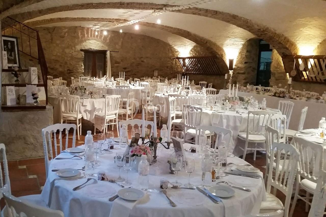 Salle voûtée en pierre aménagée pour un événement, avec des tables rondes dressées et décorées de fleurs.