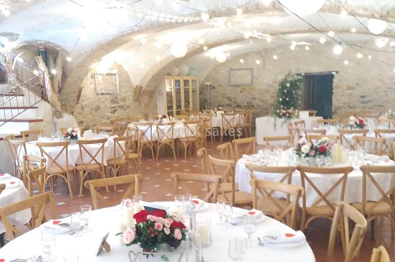 Salle voûtée décorée pour un mariage, avec tables rondes dressées, chaises en bois et éclairage chaleureux.