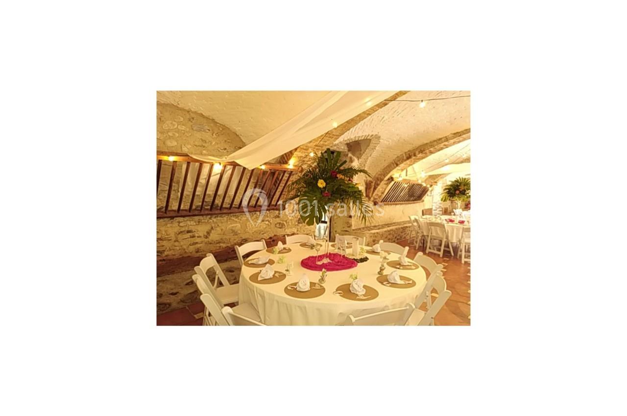 Salle voûtée décorée pour un événement, avec tables rondes dressées, nappes blanches et centres de table fleuris.