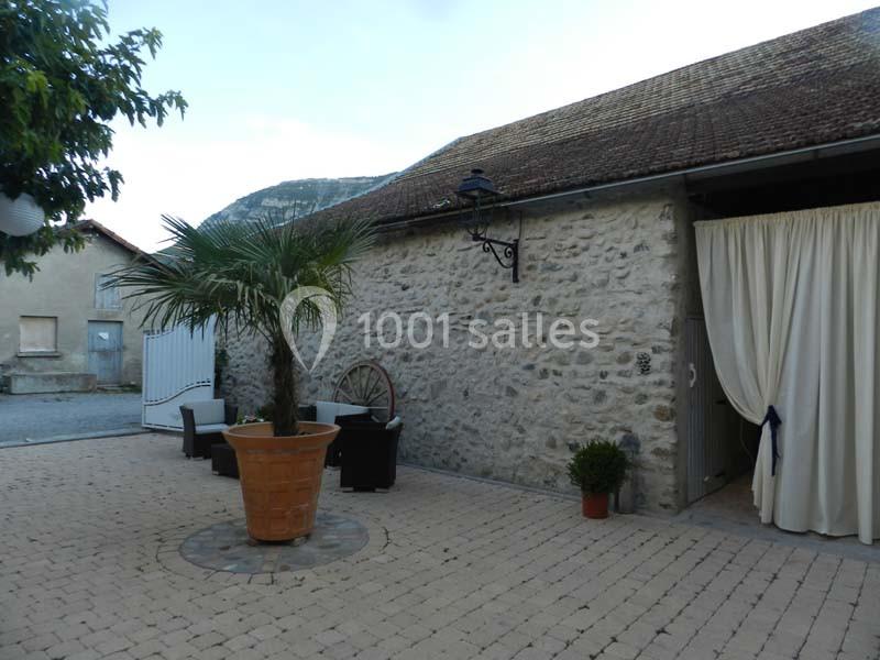 Cour pavée avec un palmier en pot, des fauteuils autour d'une table, et un bâtiment en pierre avec rideaux ouverts.
