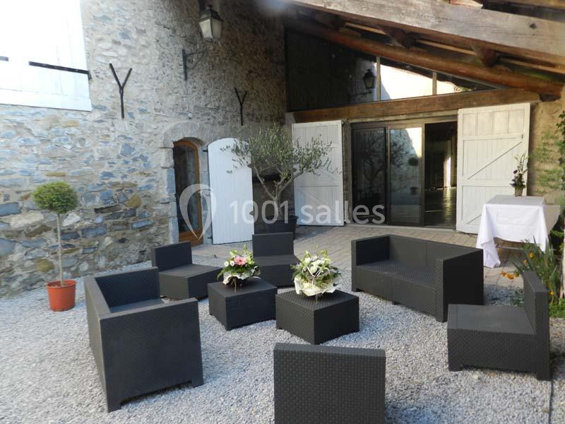 Espace extérieur avec mobilier de jardin en résine tressée, devant une bâtisse en pierre aux volets blancs.