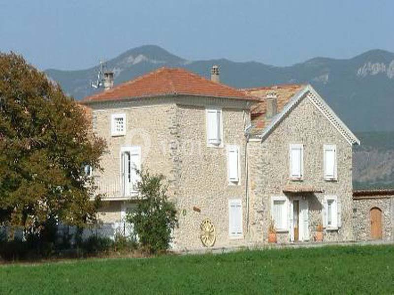 Maison en pierre avec volets blancs, toit en tuiles rouges, située dans un paysage rural avec montagnes en arrière-plan.