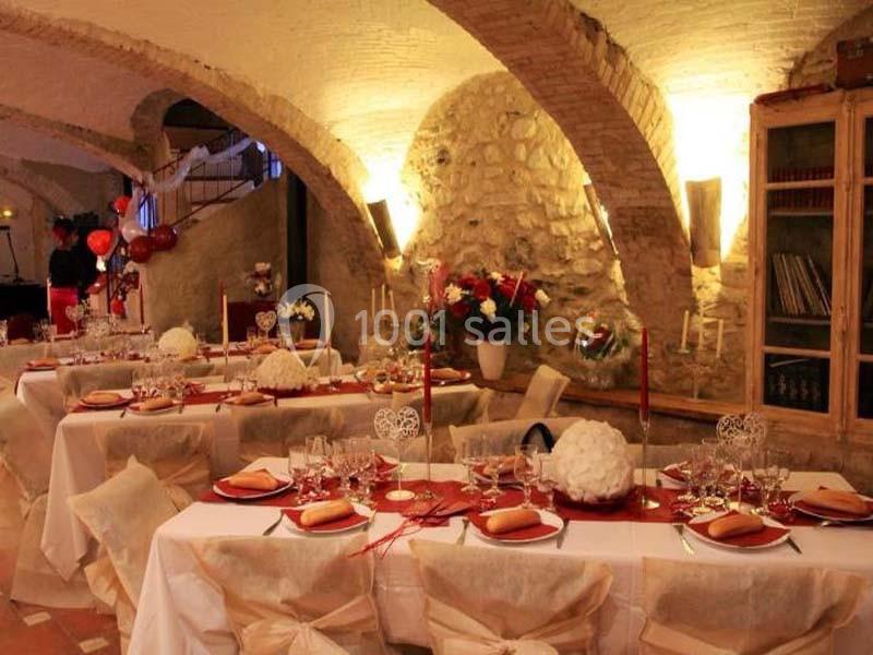 Salle voûtée décorée pour un événement, avec tables dressées, nappes blanches et ornements rouges et blancs.