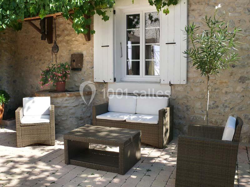 Salon de jardin en rotin avec coussins blancs, installé sur une terrasse pavée devant une maison en pierre.