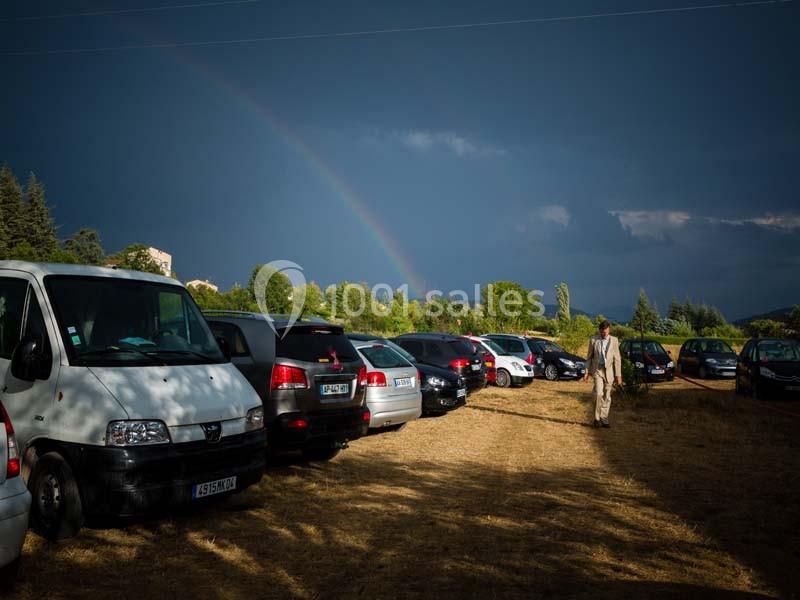 Un parking en plein air sur un terrain herbeux, avec un arc-en-ciel visible sous un ciel nuageux.