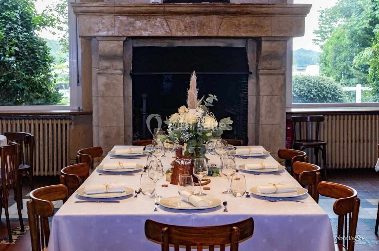 Table dressée avec une nappe blanche, vaisselle élégante et centre de table floral, devant une cheminée en pierre.