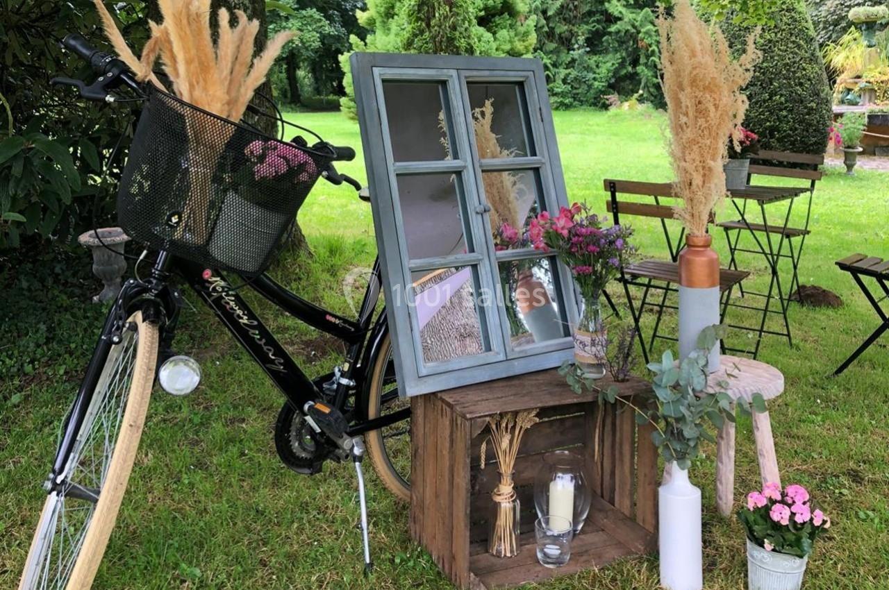 Décoration champêtre avec vélo, cadres, fleurs séchées et vases disposés dans un jardin verdoyant.