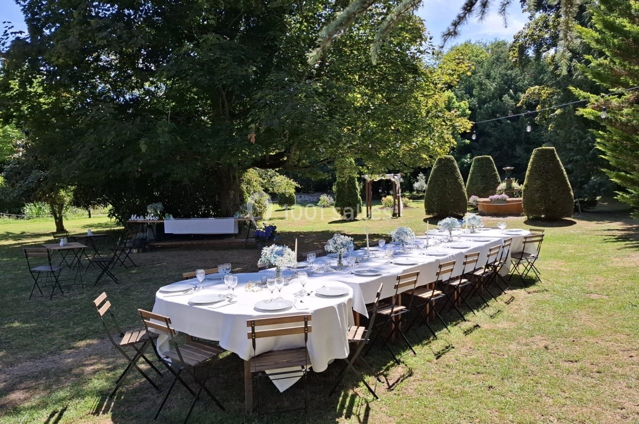 Table dressée en extérieur sous des arbres, avec nappes blanches, vaisselle et chaises en bois, dans un jardin.