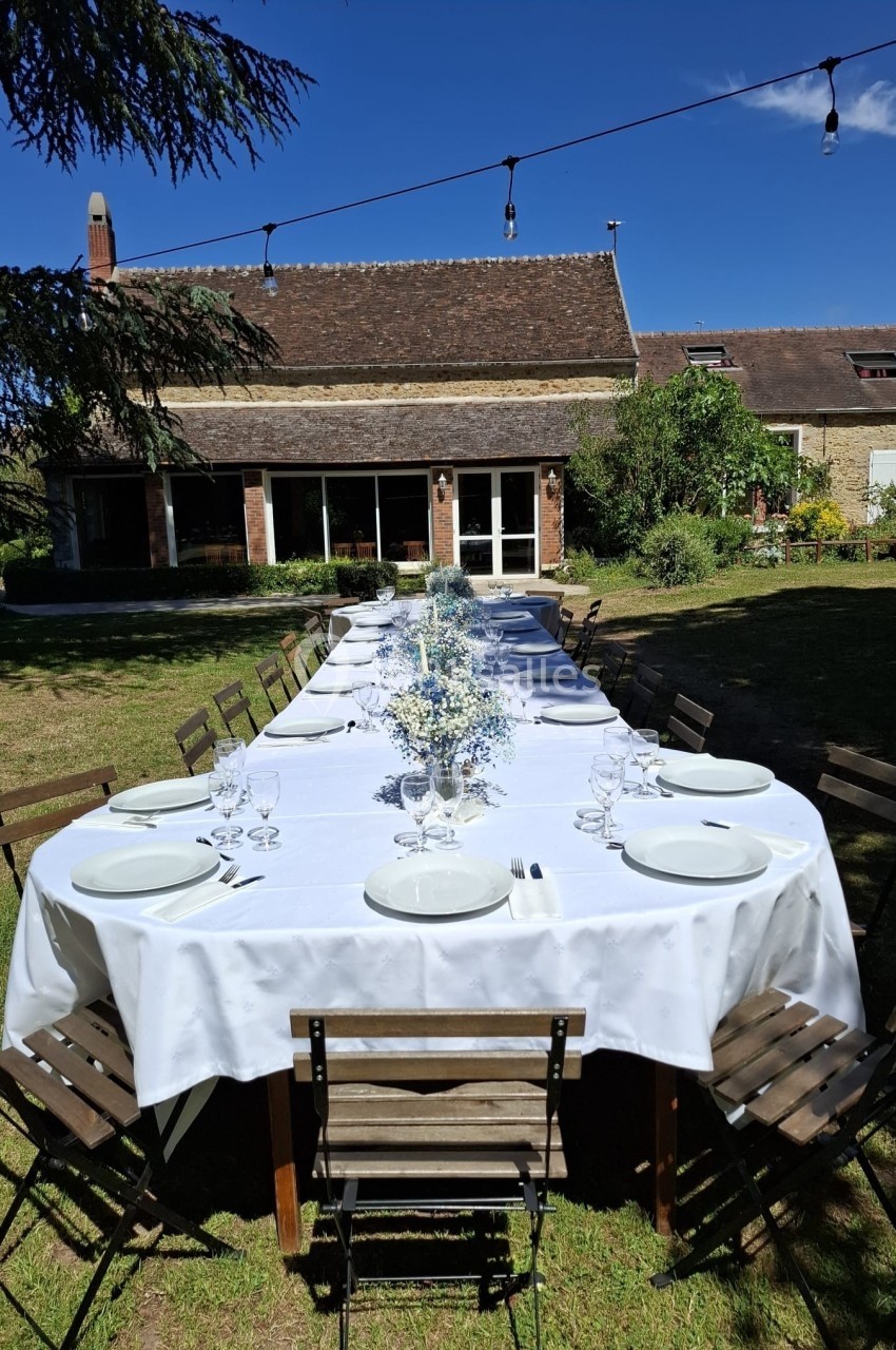 Grande table extérieure dressée avec nappes blanches, assiettes et verres, dans un jardin devant une maison en pierre.
