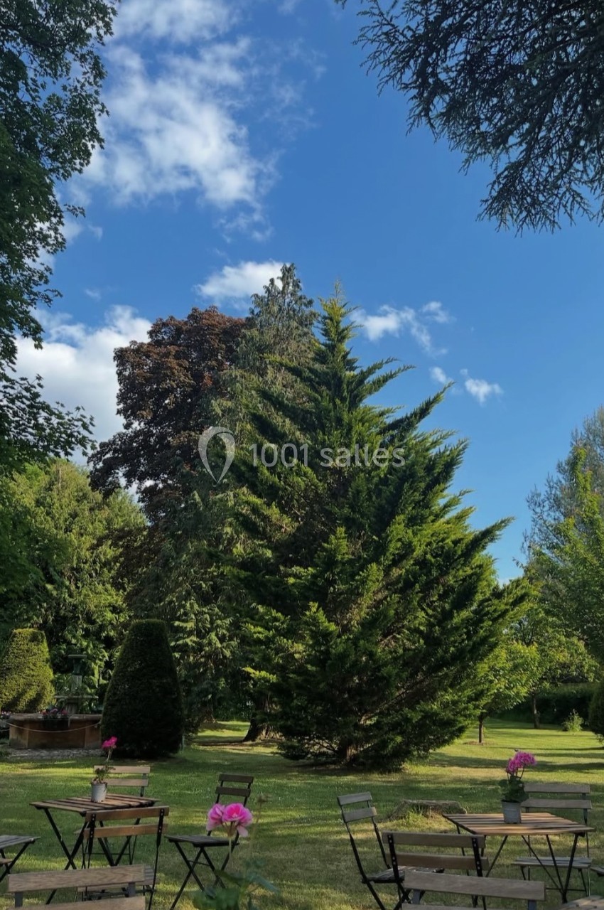 Chaises et tables en bois disposées dans un jardin verdoyant sous un ciel bleu parsemé de nuages.