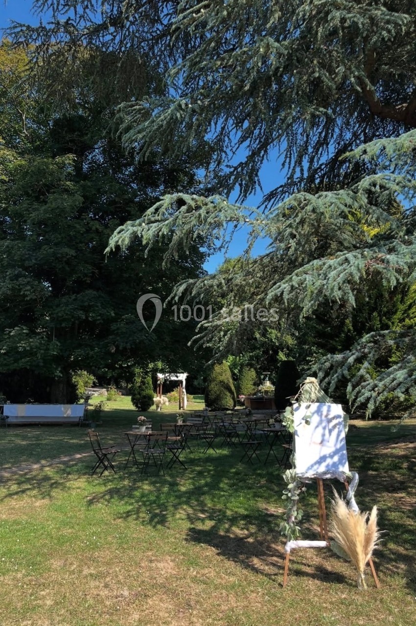 Chevalet décoré et chaises disposées dans un jardin ombragé, entouré d'arbres et de verdure.