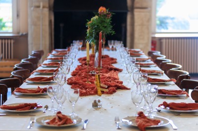 Grande table dressée avec nappes blanches, serviettes rouges, verres et couverts, décorée de bougies et fleurs au centre.
