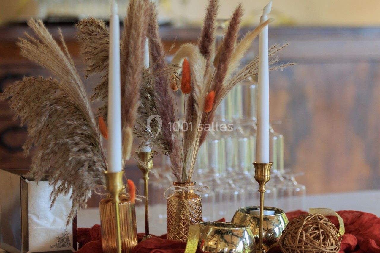 Bougies blanches et décorations dorées entourées de pampas séchées sur une table ornée de tissu rouge.