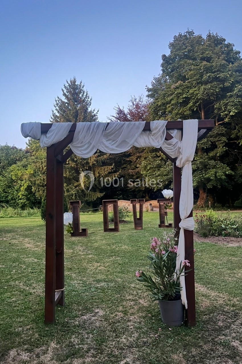 Arche en bois décorée de voilages blancs, entourée de verdure, avec le mot ’LOVE’ en lettres géantes à l'arrière-plan.