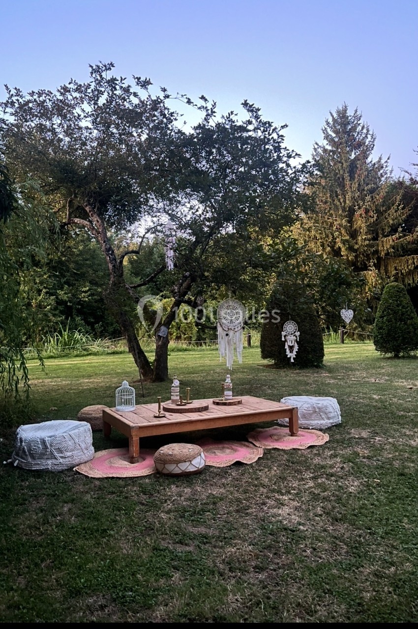 Espace extérieur aménagé avec une table basse en bois, coussins et décorations bohèmes sous un arbre dans un jardin.
