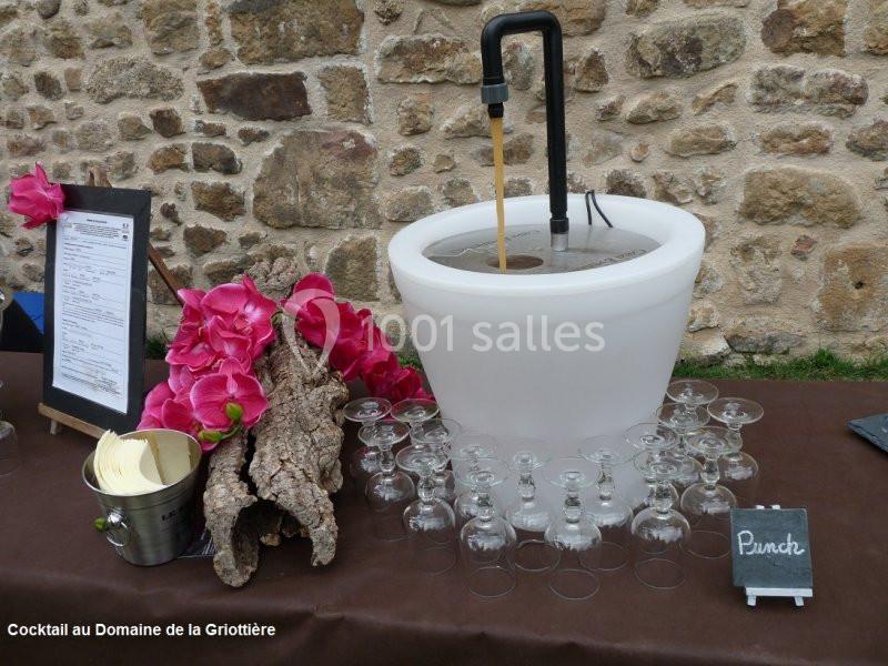 Fontaine à punch blanche avec des verres disposés autour, décorée de fleurs roses et d'écorces de bois.