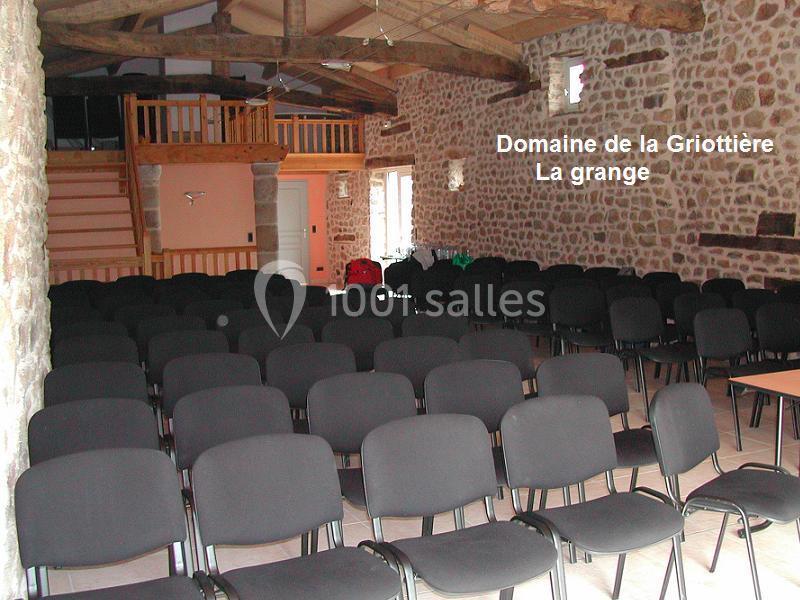 Salle en pierre avec des rangées de chaises noires disposées pour un événement, poutres apparentes au plafond.