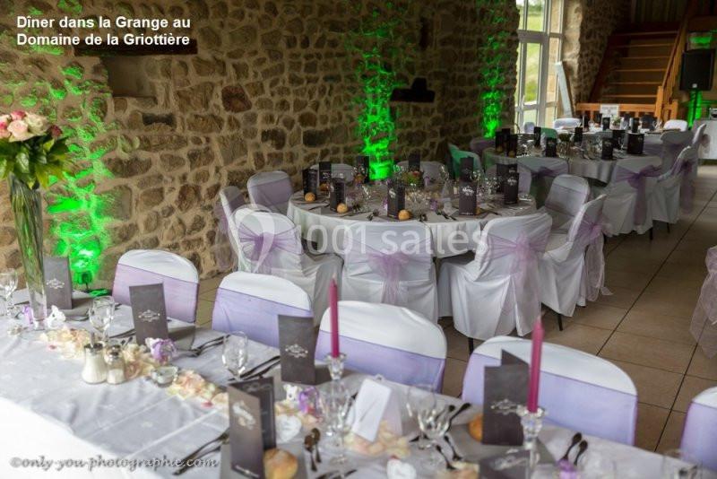 Salle de réception décorée avec des tables dressées, nappes blanches, chaises ornées de nœuds mauves et éclairage vert.
