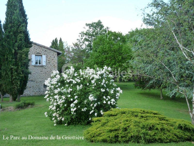 Un jardin verdoyant avec des arbres, des buissons fleuris et une maison en pierre visible en arrière-plan.