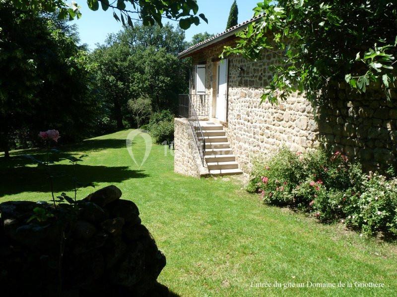 Maison en pierre avec escalier, entourée de pelouse, arbres et buissons fleuris, sous un ciel dégagé.