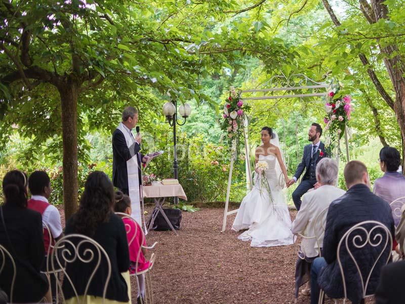 Cérémonie de mariage en plein air sous des arbres, avec un officiant, les mariés et des invités assis.