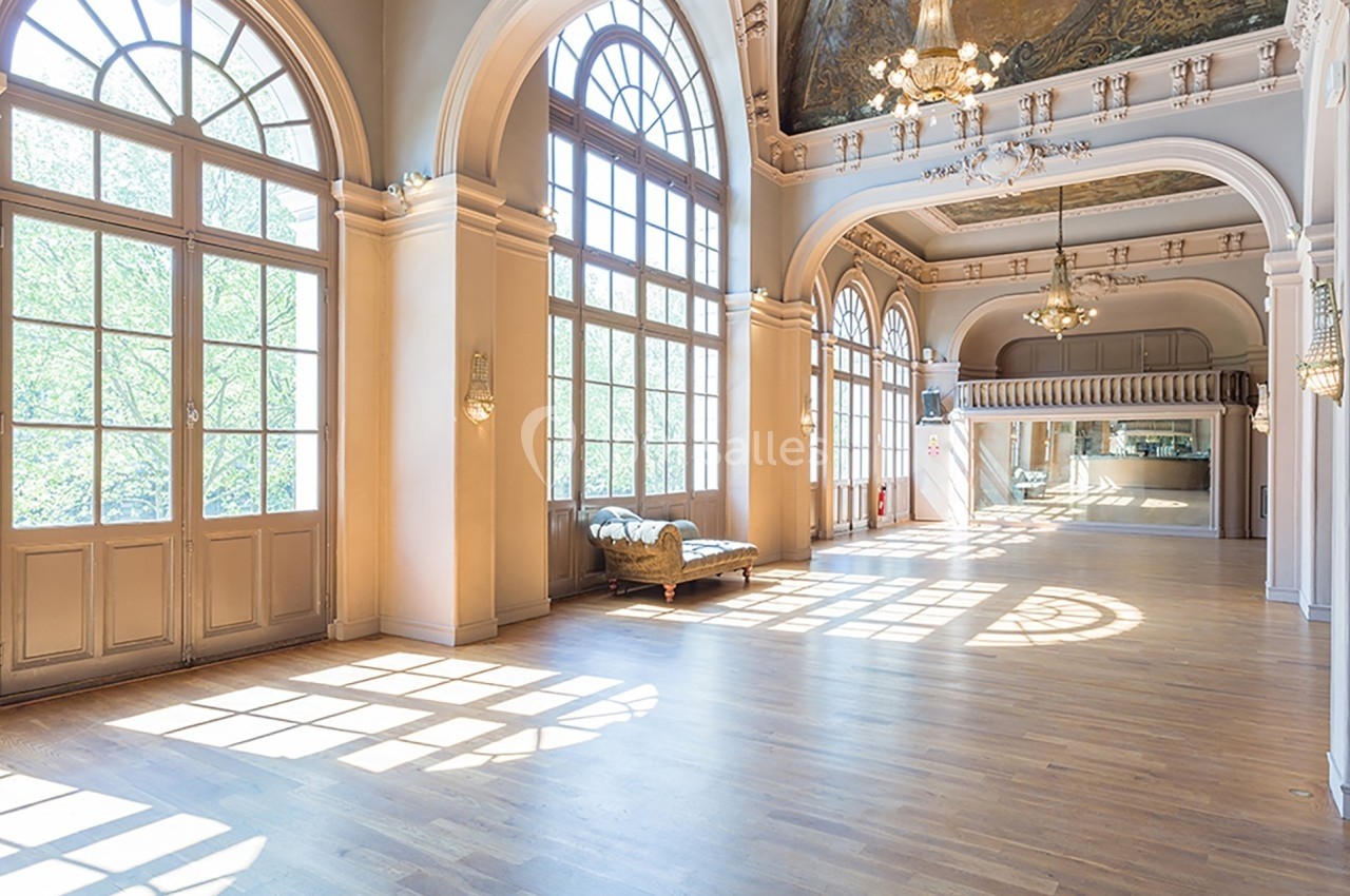 Salle lumineuse avec parquet en bois, grandes fenêtres cintrées, murs clairs et plafond orné de fresques et lustres.