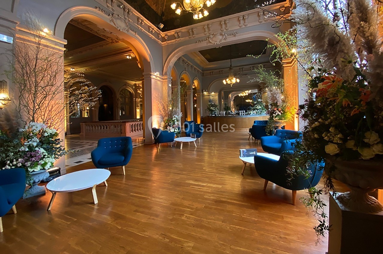 Salon élégant avec parquet, fauteuils bleus, tables basses blanches et décorations florales sous un éclairage chaleureux.