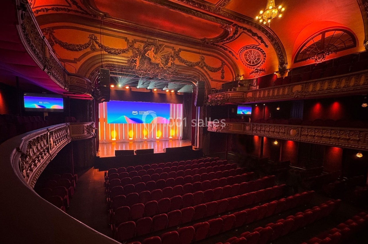 Salle de théâtre ornée avec sièges rouges, scène éclairée et écrans affichant une présentation.