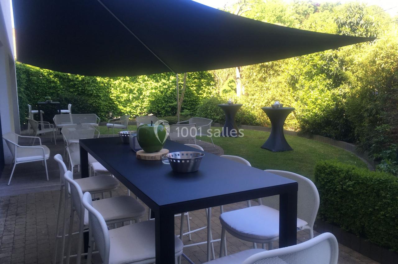 Terrasse aménagée avec table haute, chaises blanches, parasol noir et verdure en arrière-plan.