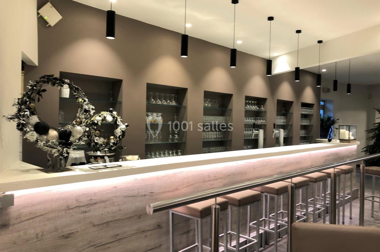 Intérieur d'un bar moderne avec comptoir éclairé, tabourets alignés et étagères remplies de verres en arrière-plan.