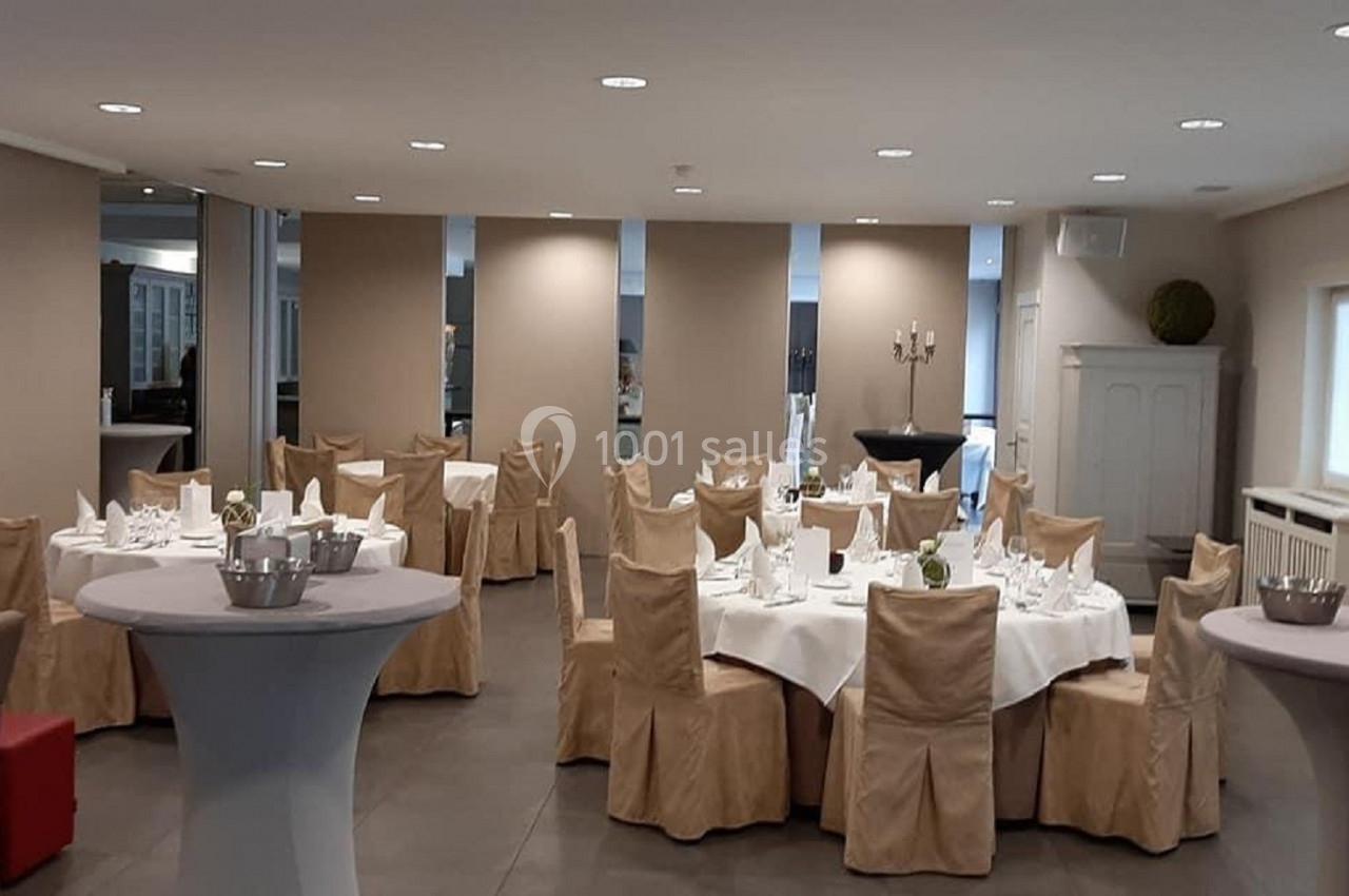 Salle de réception avec tables rondes dressées, nappes blanches et chaises couvertes, éclairée par des spots au plafond.