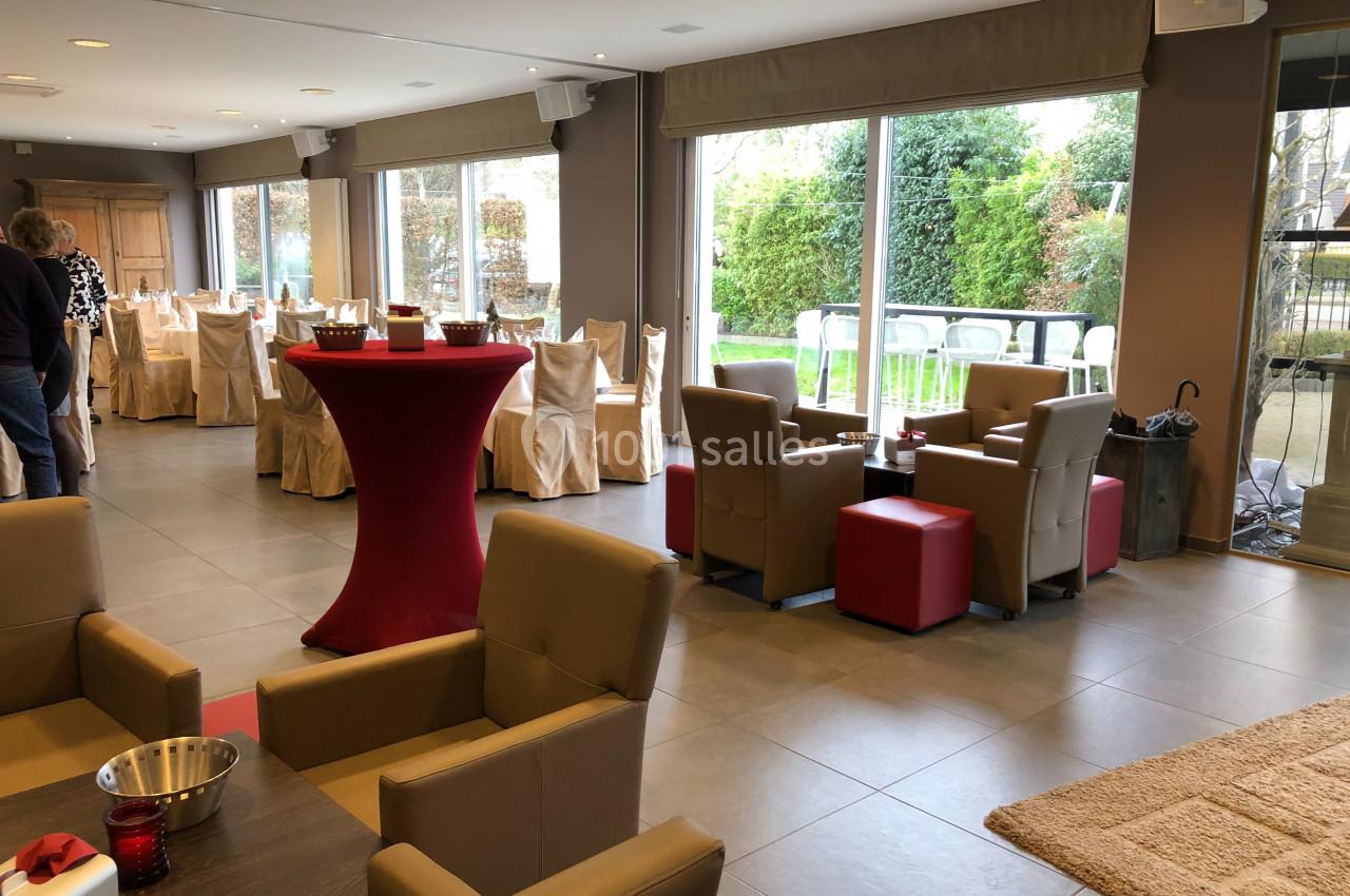 Salle lumineuse avec grandes baies vitrées, tables dressées, fauteuils beiges et tables hautes rouges.