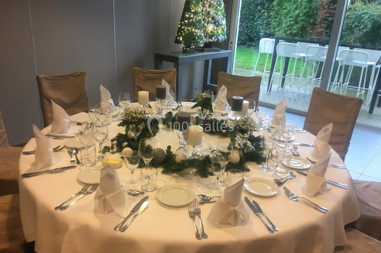 Table ronde élégamment dressée avec vaisselle, serviettes pliées et décoration florale, près d'une baie vitrée et d'un sapin…