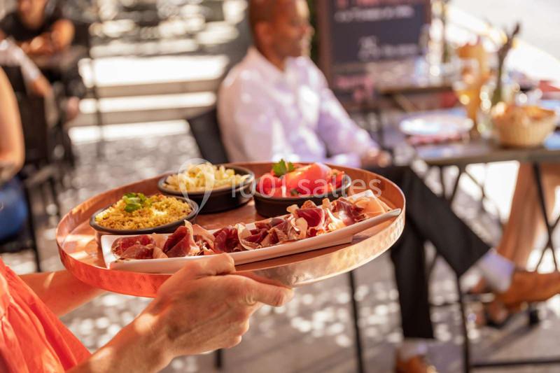 Plateau de charcuterie et accompagnements servi en terrasse, avec des clients assis en arrière-plan flou.