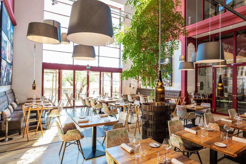Salle de restaurant lumineuse avec de grandes baies vitrées, des tables en bois, des chaises rembourrées et des suspensions…