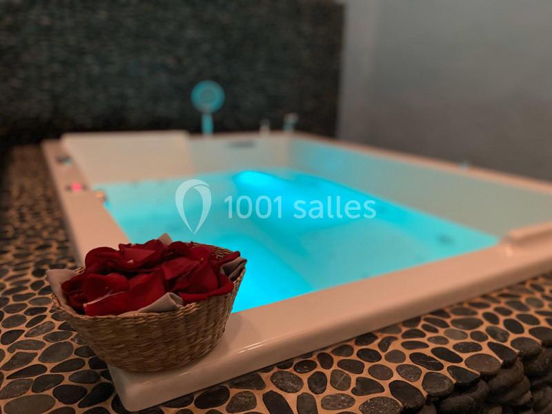 Bain à remous éclairé en bleu, entouré de pierres sombres, avec un panier de pétales rouges au premier plan.