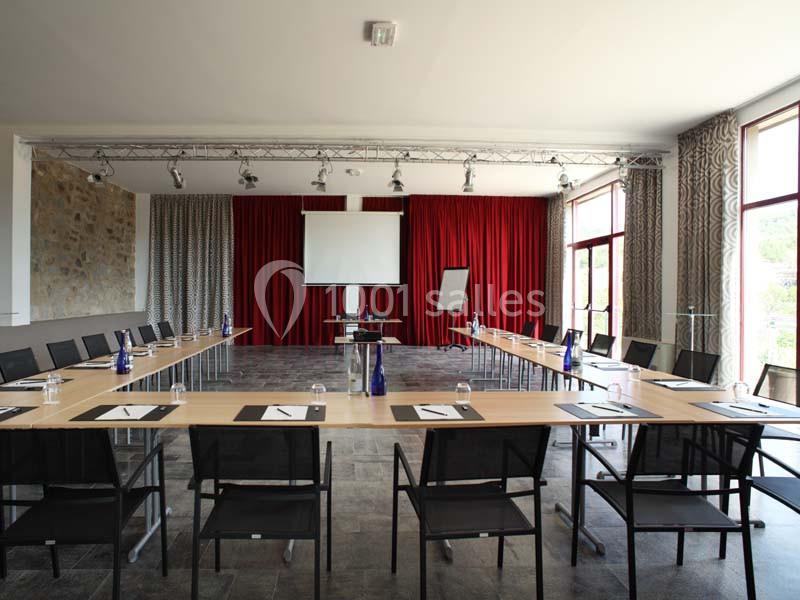 Salle de réunion lumineuse avec tables disposées en U, écran de projection, chevalet et rideaux rouges en arrière-plan.