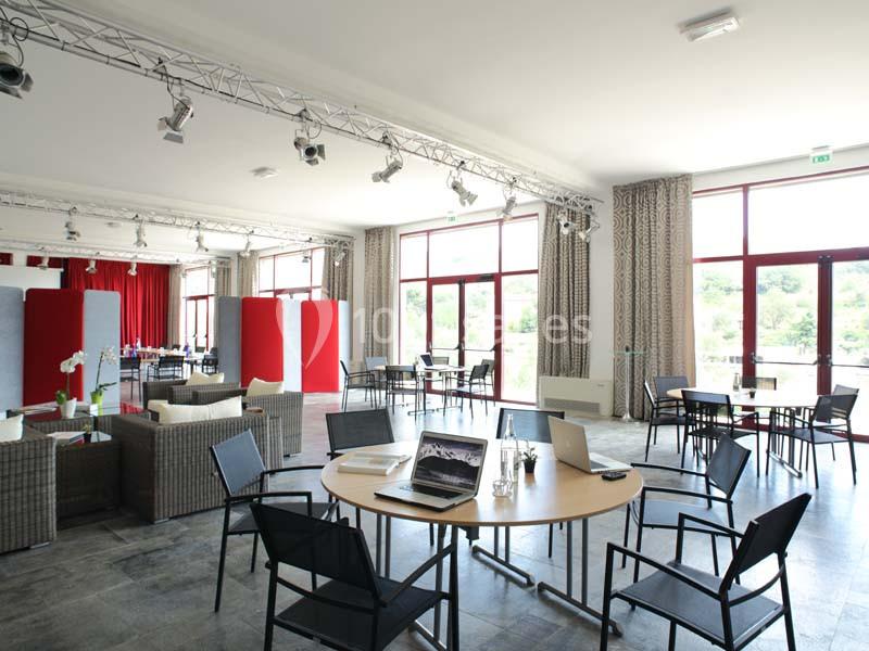 Salle lumineuse avec baies vitrées, tables rondes, chaises noires, canapés en rotin et rideaux beiges.