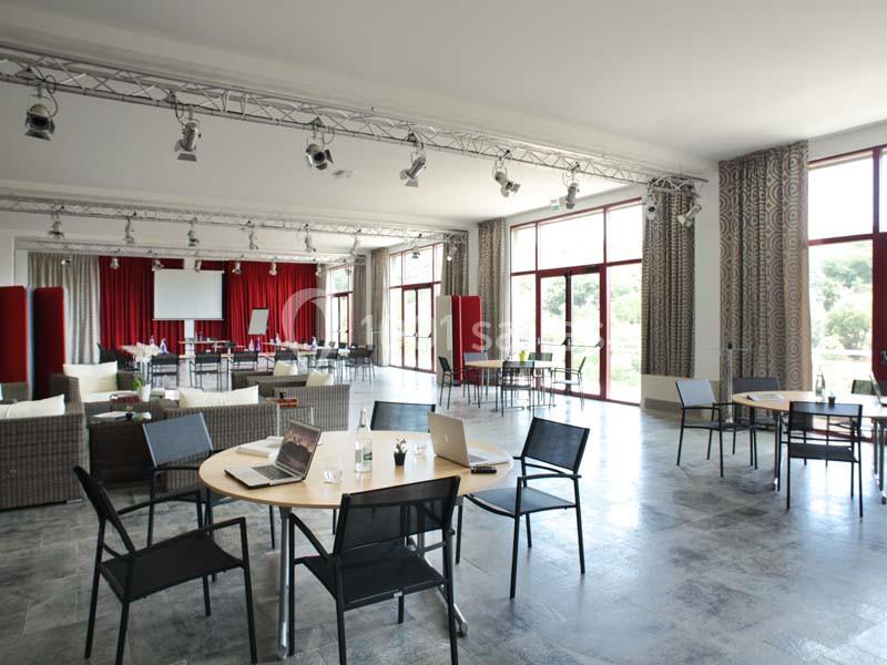 Salle lumineuse avec grandes baies vitrées, tables rondes, chaises noires, rideaux rouges et équipement de présentation.