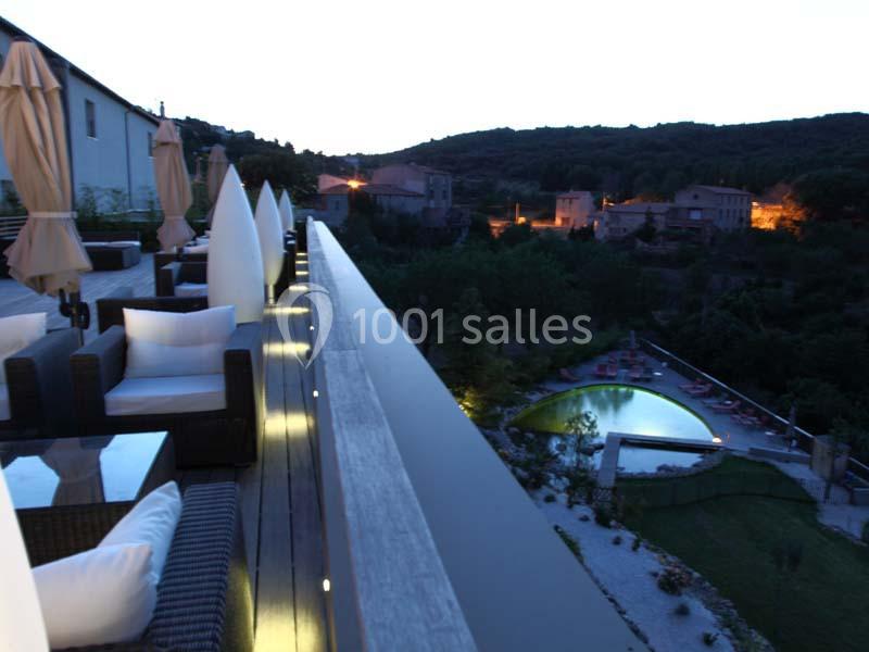 Terrasse aménagée avec canapés, vue sur un jardin éclairé et une piscine en soirée, entourés de collines et de maisons.